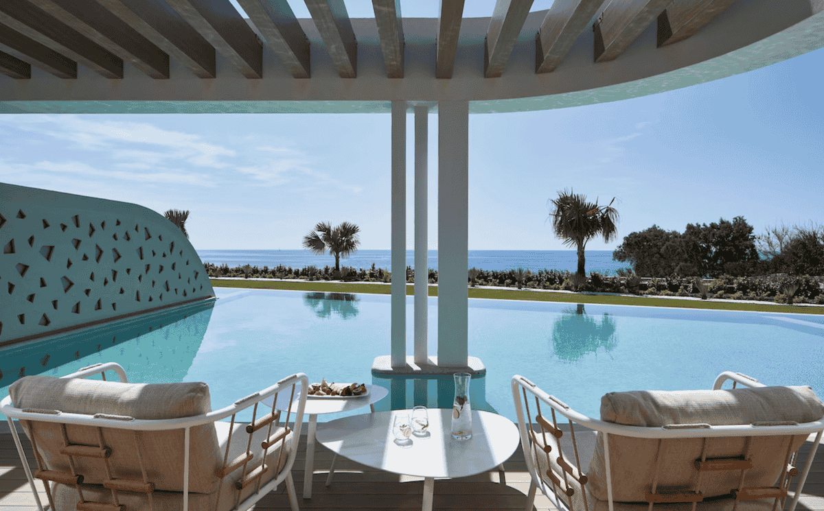A serene poolside lounge at Mayia Exclusive Resort & Spa in Rhodes, featuring modern white chairs and a table set with refreshments. The infinity pool seamlessly blends with the horizon, offering breathtaking ocean views framed by palm trees and minimalist architecture.