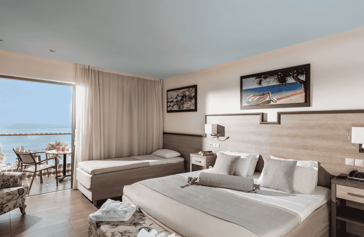 A bright and inviting hotel room at Star Beach Village and Water Park, featuring a comfortable double bed, a separate daybed, and a private balcony with a sea view. Neutral tones, soft lighting, and beach-themed artwork create a relaxing coastal ambiance.
