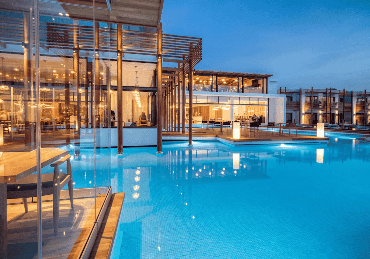 A stunning evening view of Stella Island Resort & Spa, featuring a luxurious overwater dining area surrounded by a glowing blue pool. The modern architecture, warm ambient lighting, and sleek wooden pergolas create an elegant and tranquil atmosphere for guests.