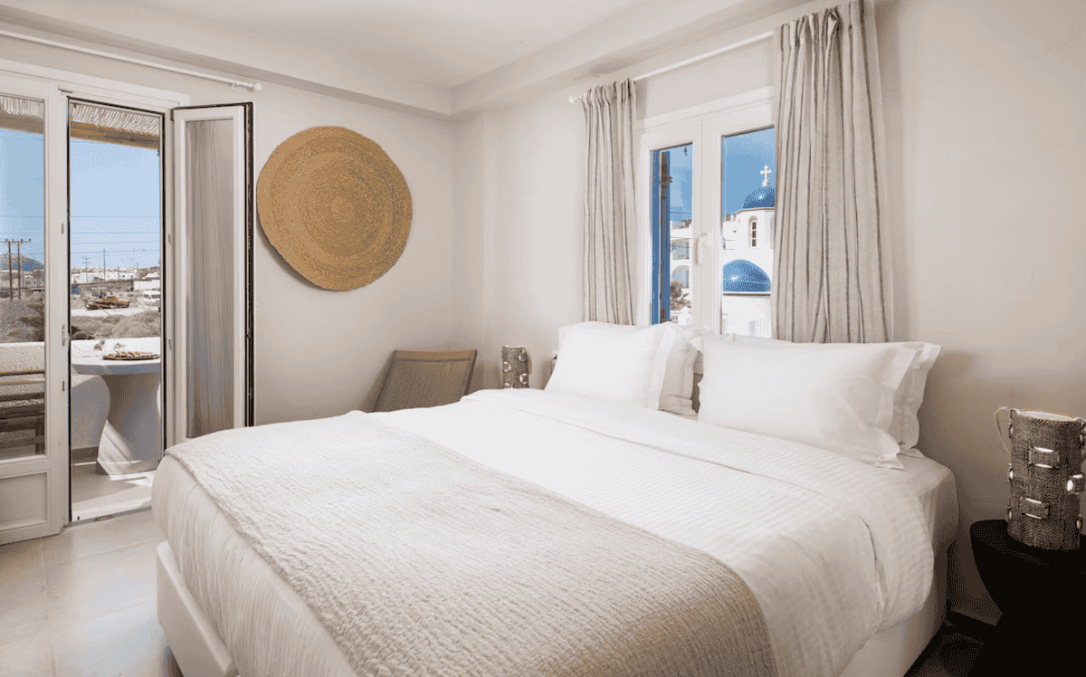 A bright and airy bedroom at Noverian Bios Santorini, a vegan and vegetarian boutique hotel. The room features a plush white bed, neutral decor, and large windows offering a stunning view of Santorini’s iconic blue-domed churches and a private balcony with outdoor seating.